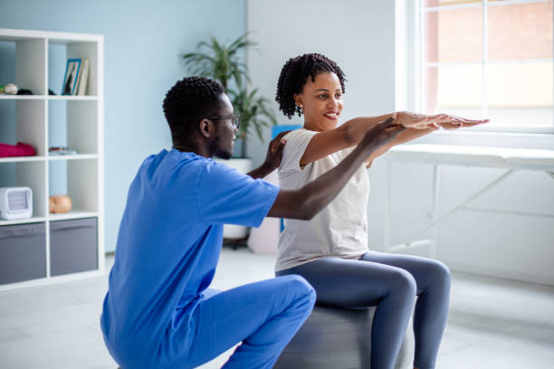 A physiotherapist assists a smiling woman balancing on a gray exercise ball, focusing on arm extension exercises in a bright, modern therapy room setting.